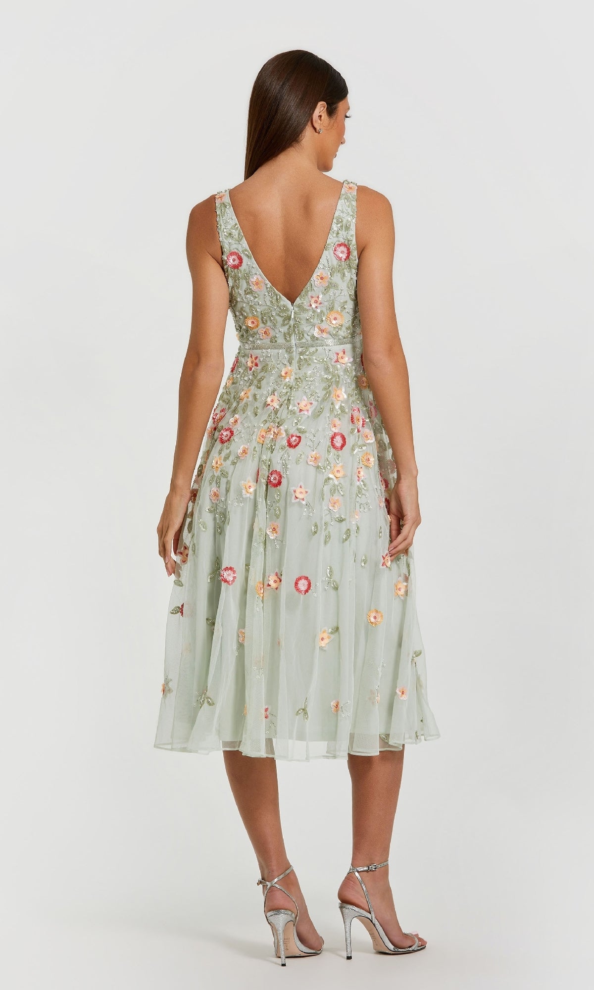 A woman faces away, wearing the Mac Duggal 5527 floral-embroidered party dress in light green, sleeveless and tea-length, paired with silver high-heeled sandals.